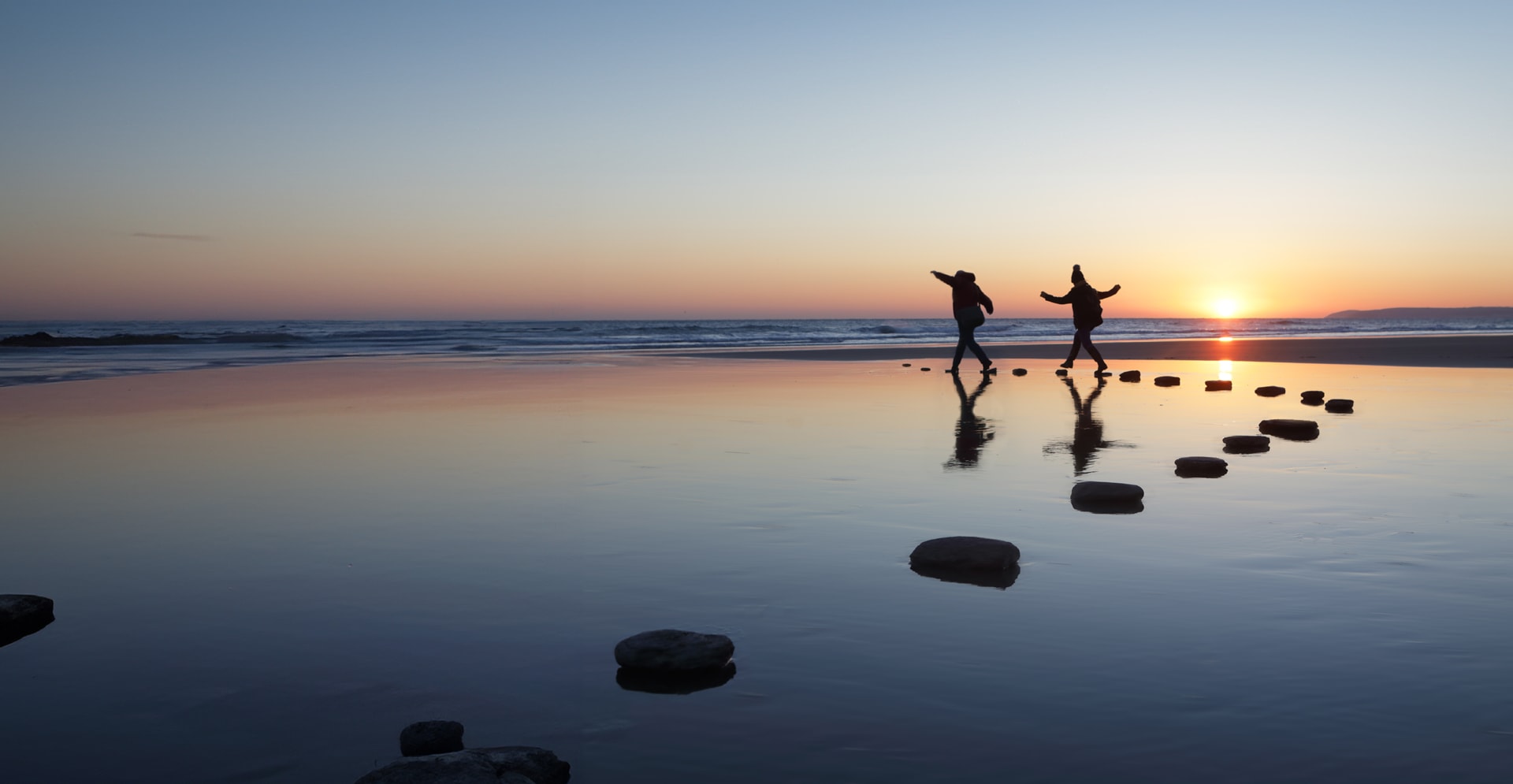 Two individuals skipping on rocks along the beach, silhouetted against a vibrant sunset sky.