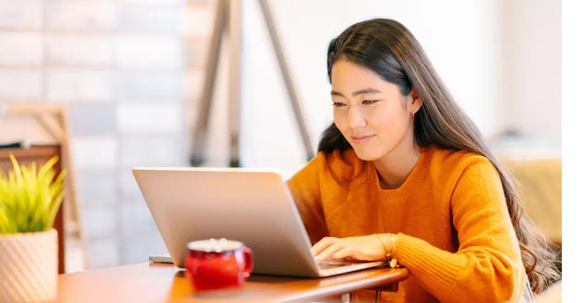 Woman in an orange sweater sits at a table, working on a laptop