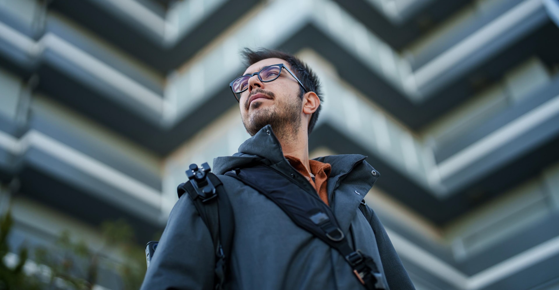 A man in glasses and a jacket poses in front of a building, showcasing a professional demeanor.