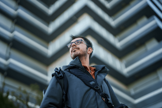 A man in glasses and a jacket poses in front of a building, showcasing a professional demeanor.