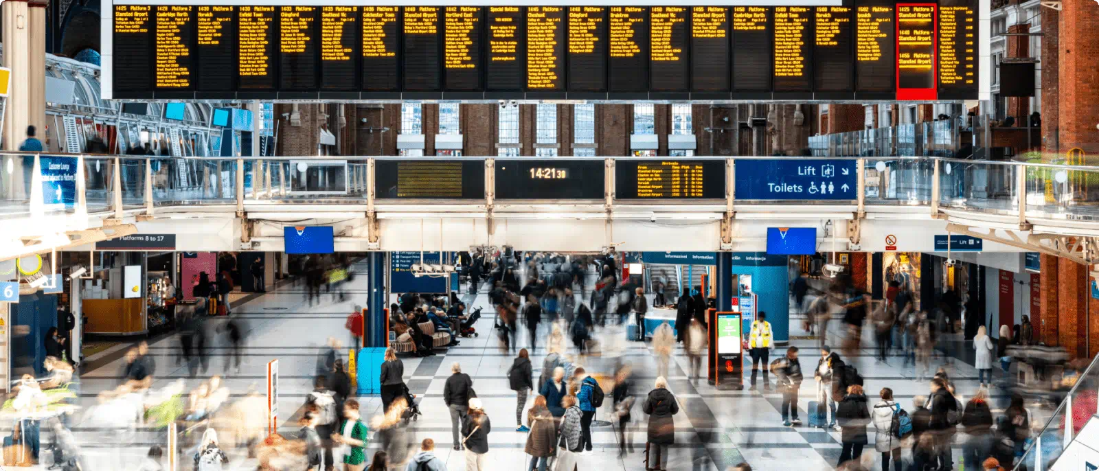 People walking through a busy train station with a directional sign visible on the wall.